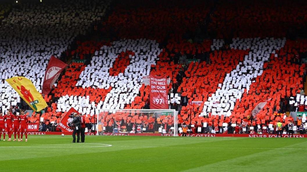 Fans in a packed stand at Anfield stadium display the number 97 against a red backdrop while players stand for a minute's silence on the pitch