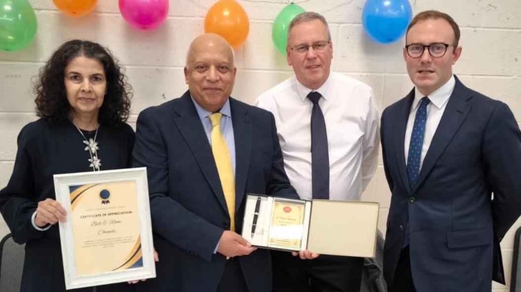 A woman and three men standing in front of a white wall with balloons hanging from the top. The woman on the left is holding a framed certificate and the man next to her is holding a pen in a box.