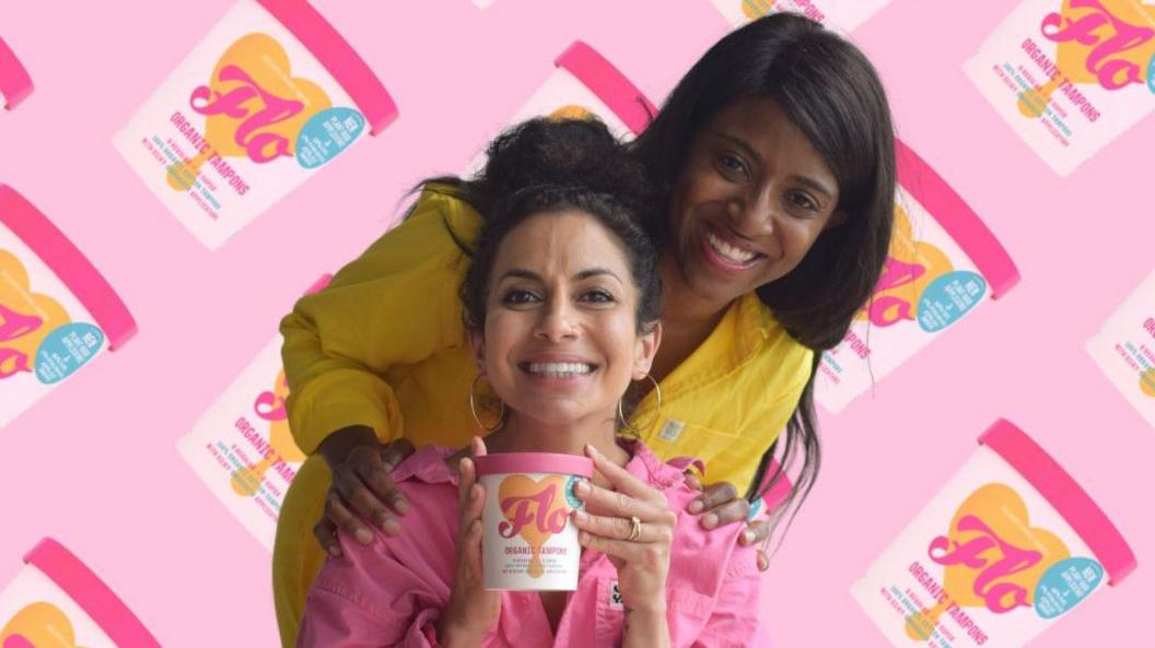 Tara Chandra and Susan Allen. Tara is pictured in the centre of the frame wearing a pink shirt and holding a tub of Flo tampons. She is looking directly at the camera and smiling. Susan is stood behind Tara, with her hands on both her shoulders. She is crouched slightly with her head to the side, next to Tara's. She is wearing yellow and is also looking at the camera and smiling. The photo is edited with a pink background and a graphic pattern showing the Flo product. 