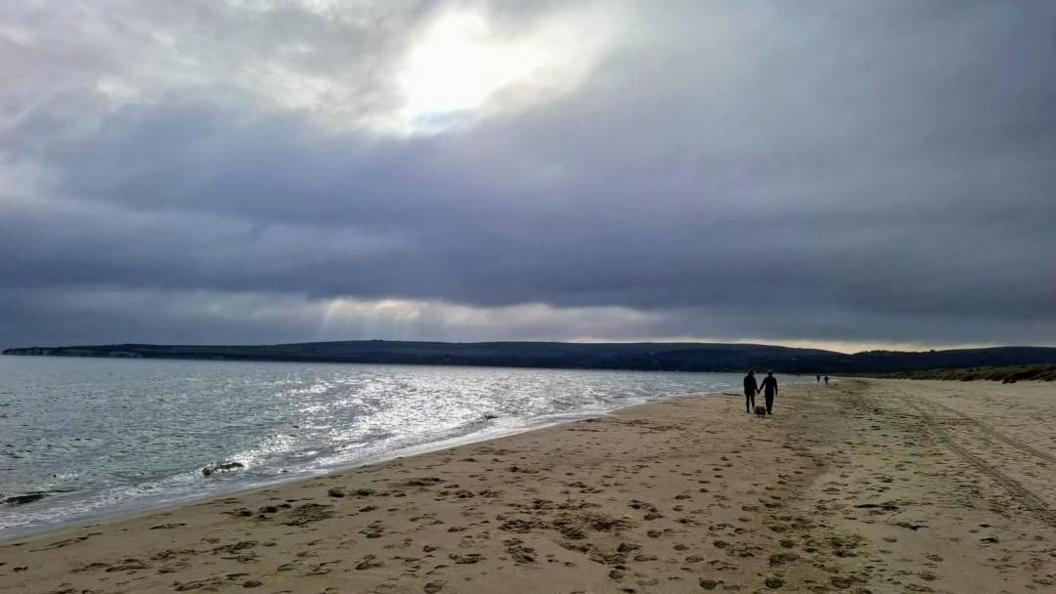 A beach scene with a threatening dary grey sky. A small number of people can be seen walking on the sand in the distance - one couple is hand-in-hand with a dog.