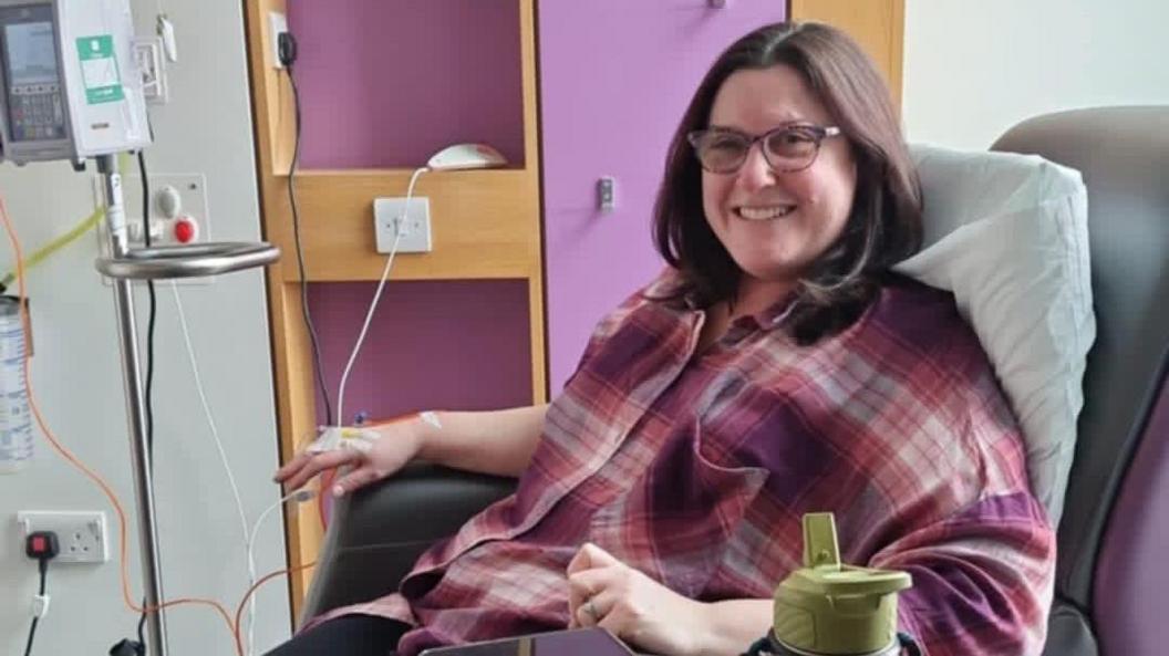 A woman with shoulder-length brown hair and glasses is smiling and sitting in a chair. She has a cannula in her hand which is wired into a machine. She is wearing a pink, purple and white plaid top.