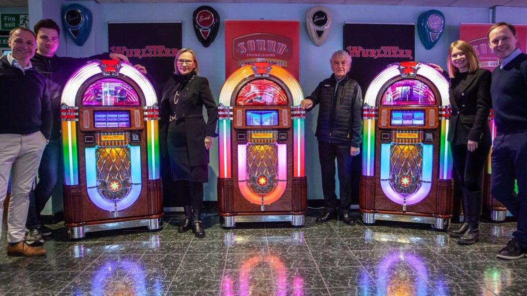 Two women and four men stand around three Wurlitzer jukeboxes lit up in bright colours.