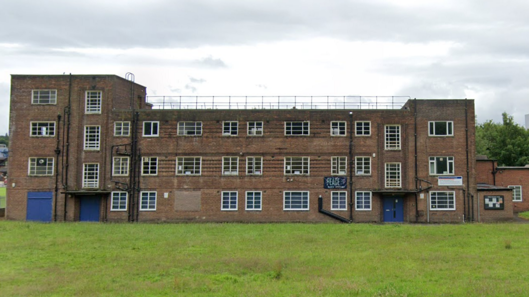 A red-brick building with three storeys and a four-storey tower at one end. It has rows of white-framed windows with one boarded up.