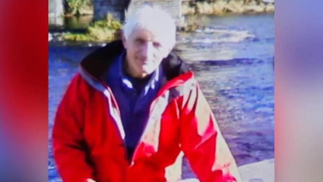 Brian Perry standing by a river, wearing a red waterproof jacket and a blue jumper. He has grey/white hair and smiles at the camera