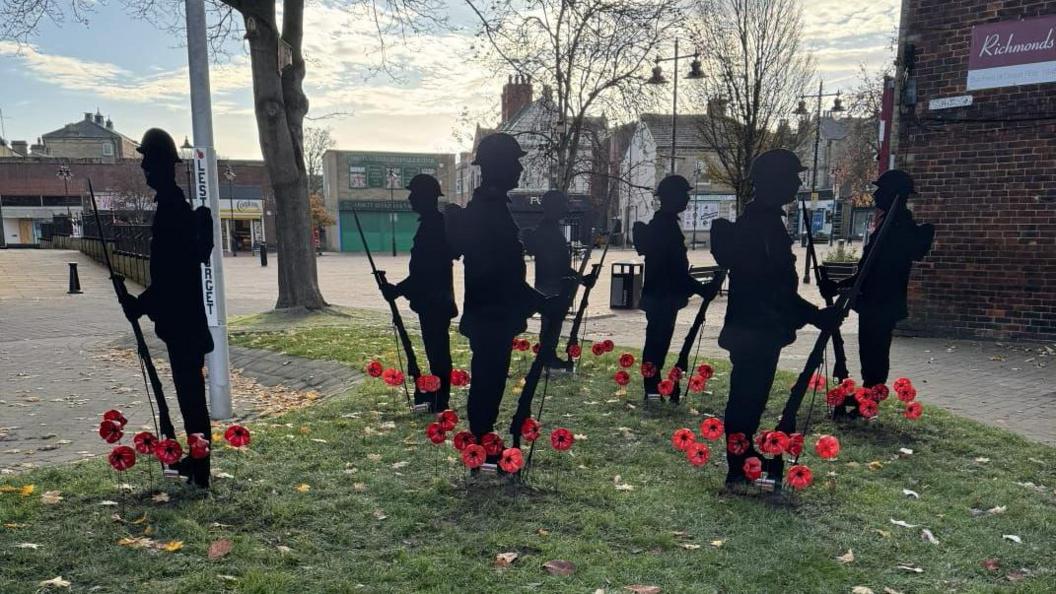 Several black silhouette figures of soldiers holding rifles are arranged on a grassy area, each surrounded by red poppy flowers. In the background, there is a brick building on the right, a large leafless tree, and other buildings further away.