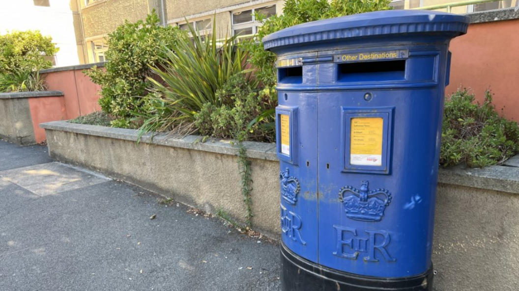 A blue double post box on a pavement. It is in front of a small wall which has plants on the other side of it. The blue post box has two yellow signs on it, and has a crown engraved at the bottom of it with the letters 'E and R' below it.