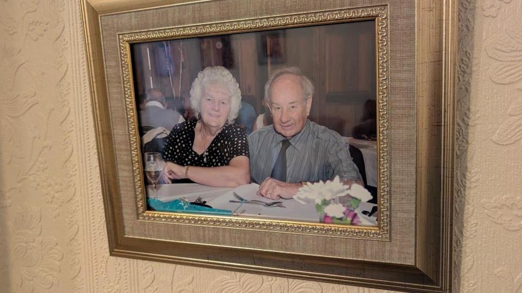 A framed picture hanging on the wall of a man and woman. The woman, on the left has short white hair and is wearing a black and white top. The man has short grey hair and is wearing a silver coloured shirt and dark grey tie. 