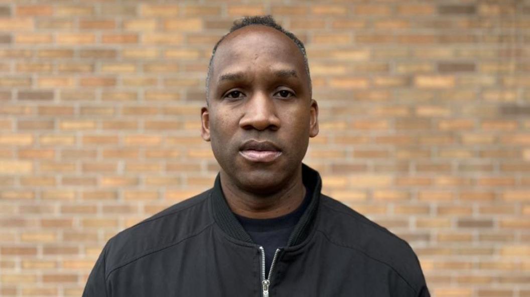 A man with short dark hair and a black zip-up jacket is standing in front of a brick wall.