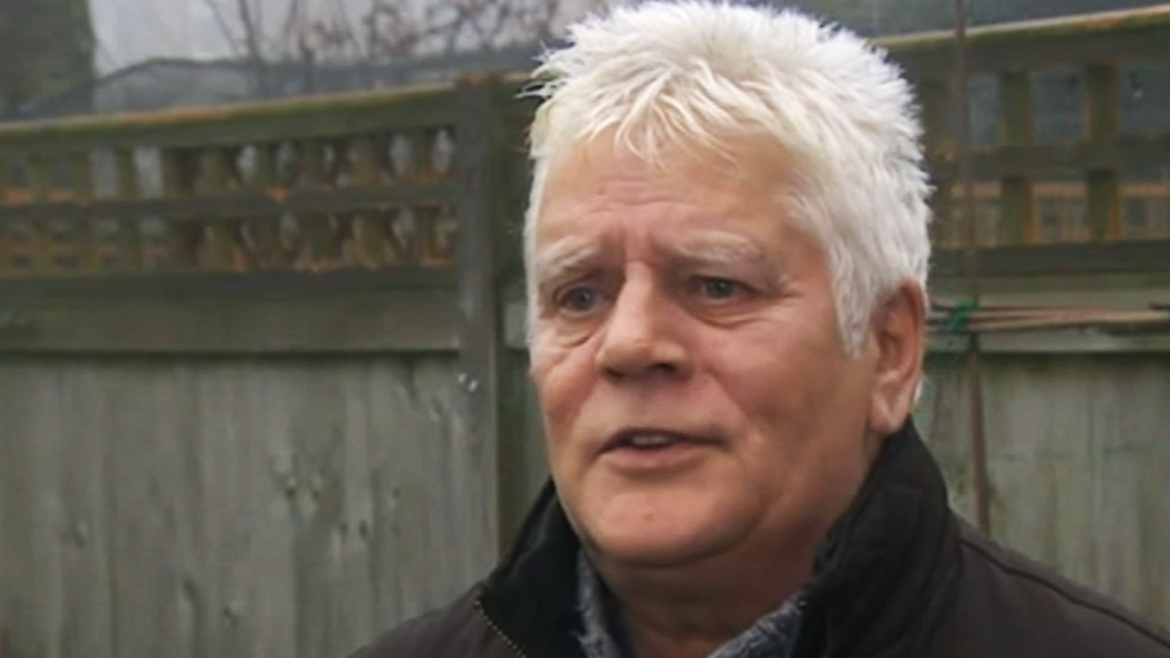 Colin Knox, a white haired man, stands in front of a wooden fence.