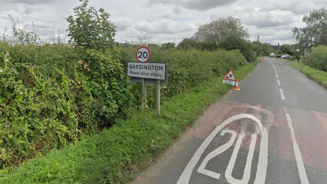 A road view of Oxford Road into Garsington. There is a sign with a 20mph speed limit. There is a roadworks sign and a cone on the road/
