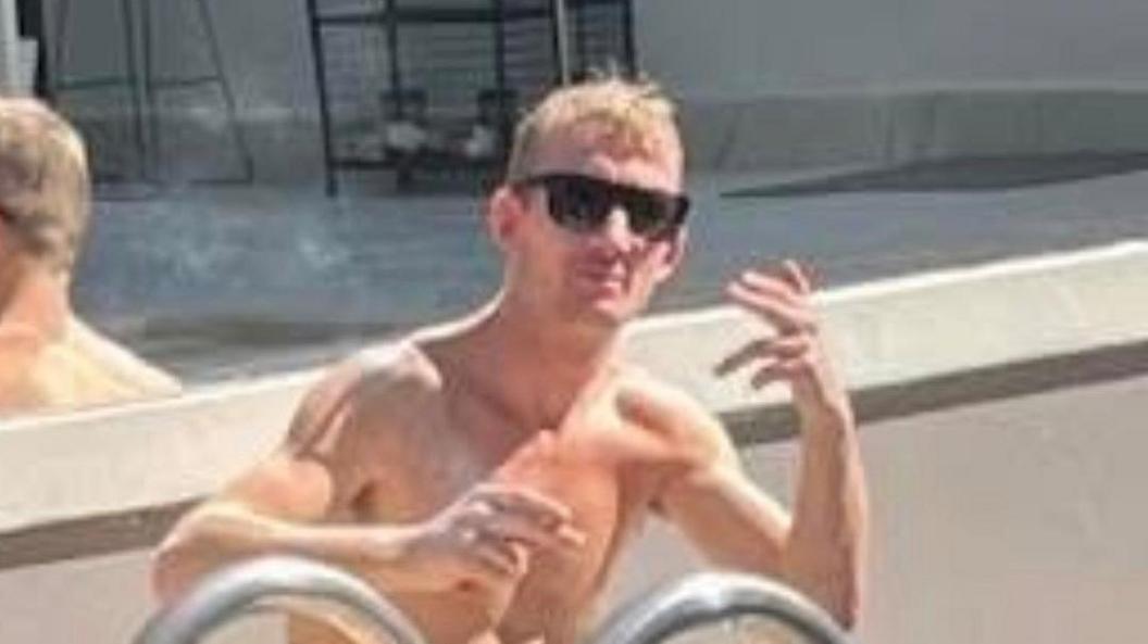 Craig Yorke is sitting, shirtless, by a pool and posing for the camera. He has short blonde hair and is wearing glasses.