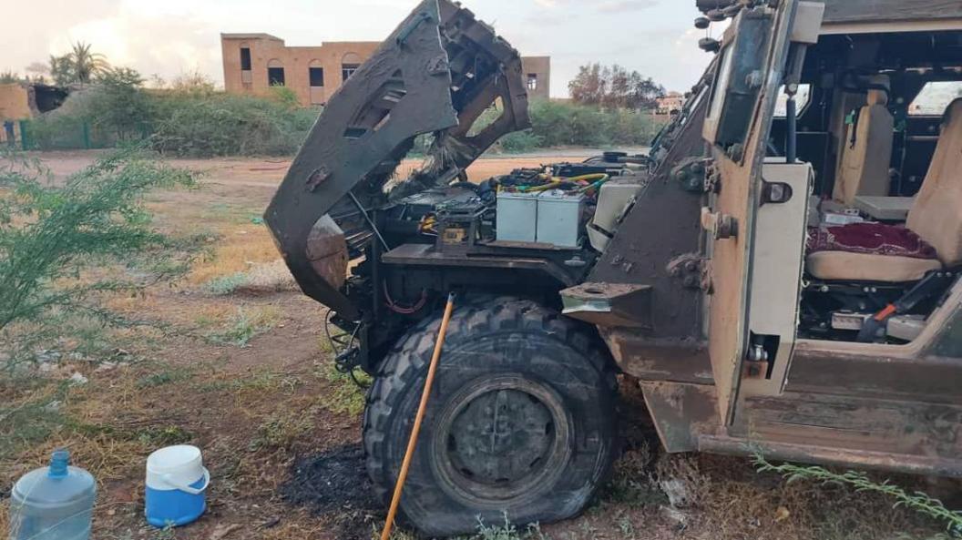 An armoured vehicle that is partially damaged with its bonnet open, with a building in the background