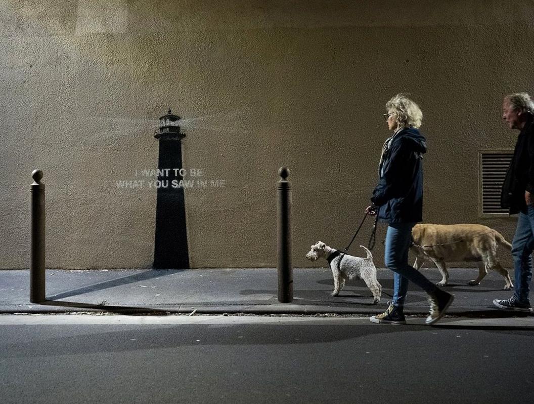 A photo of the Banksy piece of art with two dogwalkers walking past on the street. A metal bollard is in the foreground on the pavement, while a painting of a lighthouse is on the wall superimposed with a quote "I want to be what you saw in me".