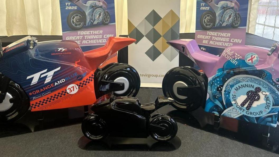 Three models of the racing bike sculptures. An orange and blue bike with the TT logo is on the left, a small black bike is in the middle, and a purple and blue bike with Mannin Cancers Support Group's logo is on the right.