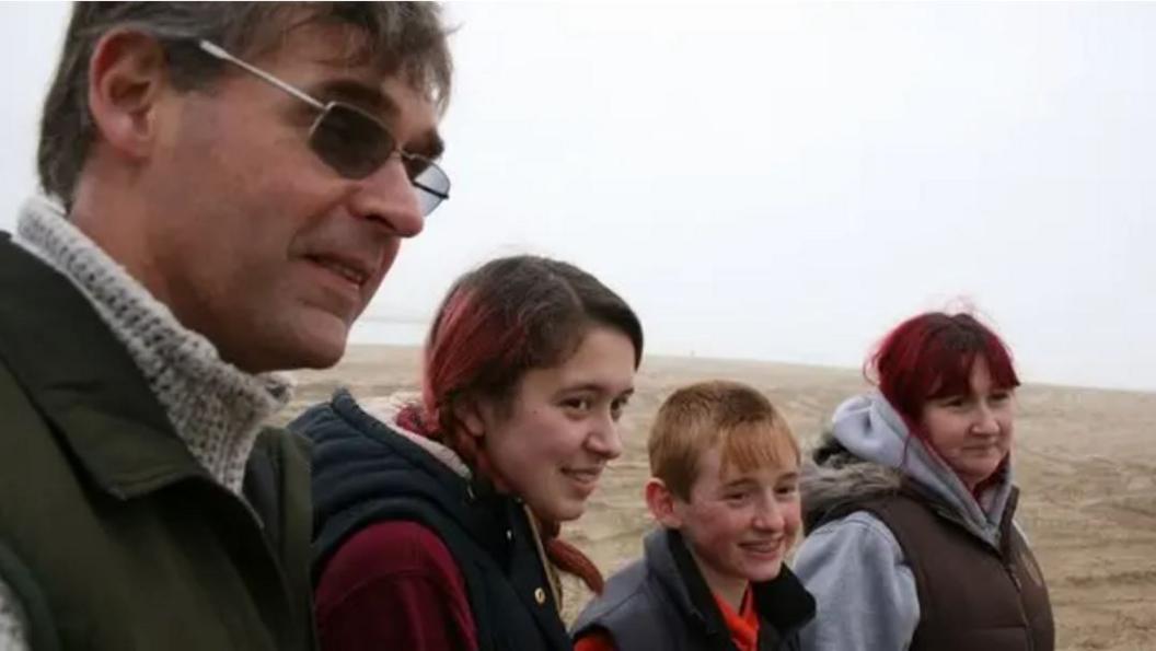 Paul Jones on the left has dark hair and wears dark glasses, a a light coloured roll-neck jumper under a dark coat. To his right is a young girl with long brown hair and a dark hooded body-warmer and red jumper. To her right is a young boy with fair hair and dark body-warmer and red top. To his right is a woman with long dark hair, a green top under a dark body-warmer.