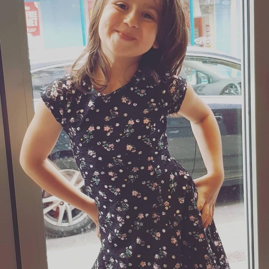 Young girl Sara Sharif stands smiling with her hands on her hips. Her hair is just past her shoulders. She is wearing a navy blue short-sleeved dress with little pink flowers on it.