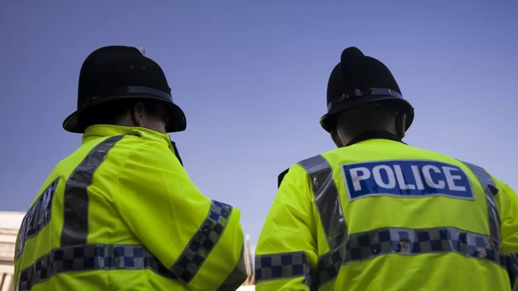 The backs of two police officers in front of a blue sky. They are wearing neon yellow jackets that say "police" and black police hats