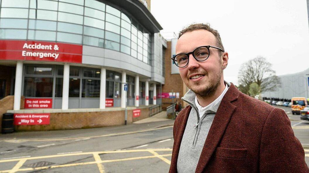 Adam Dance, smiling, in front of the Yeovil Hospital A&E