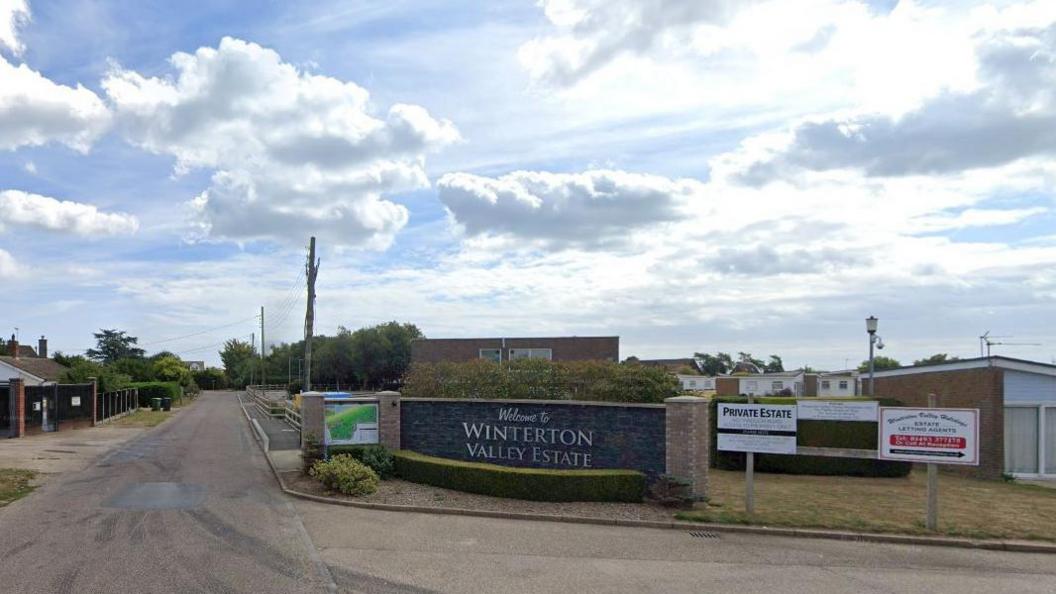 A picture of the Winterton Valley Estate holiday park entrance on the Norfolk coast by a quiet road and beneath a blue, cloudy sky.