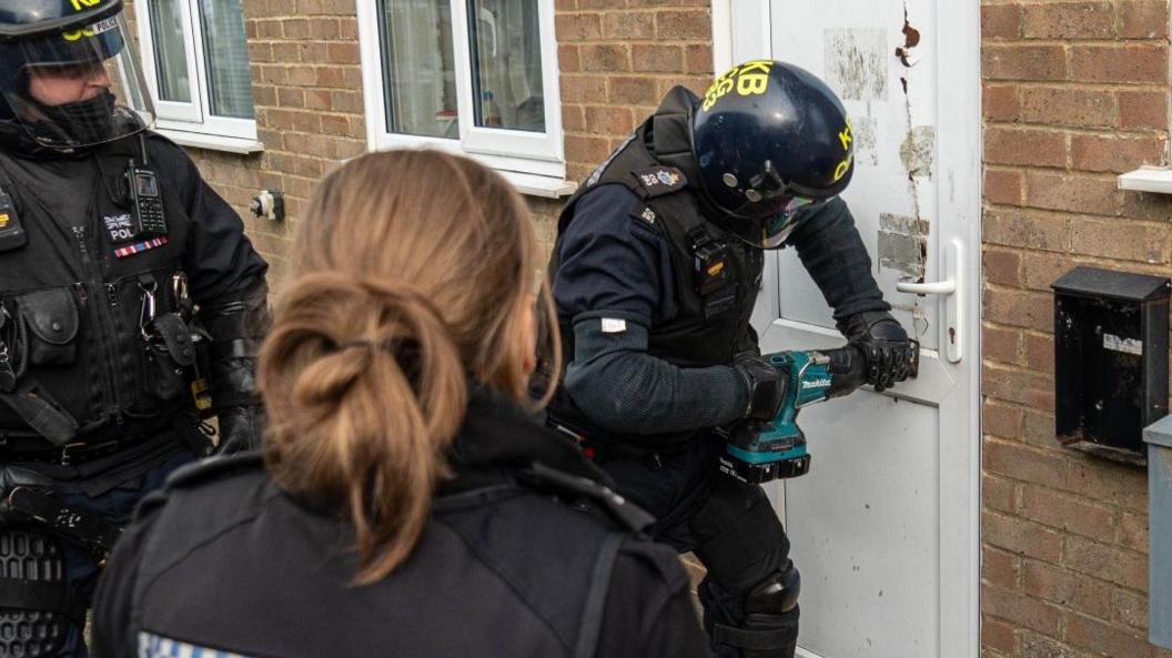 Three police officers wearing tactical uniforms break into a house, using a drill to remove the lock.