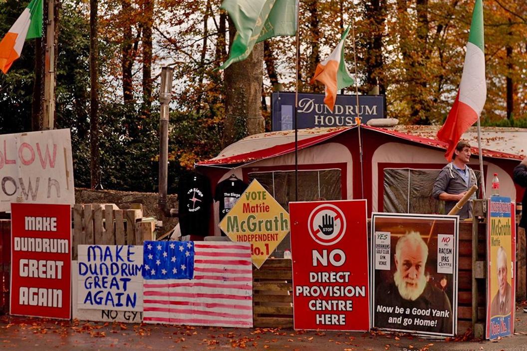 A caravan parked by protesters outside Dundrum House, with Irish flags and a variety of posters outside - some handmade, others printed - as well as an independent candidate's election sign. The posters read: "Make Dundrum great again", "No direct provision centre here", and "Now be a good yank and go home"