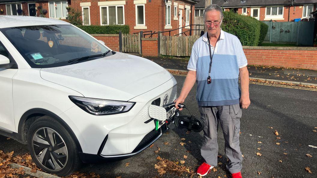 Mr Kelsall stands holding an EV charger in his white Tesla car outside his home on an autumn day - the EV charger is not attached to anything 