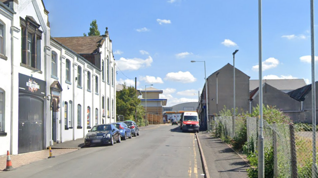 A number of parked cars are parked along the side of a street with white buildings on one side and a metal fence on the other.