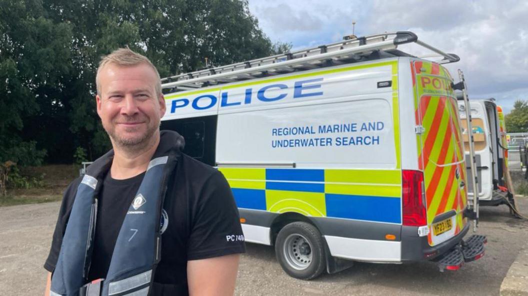 PC Liam Whittington wears a black t-shirt and blue lifejacket. He is smiling in front of a regional marine and underwater search police van, which is white with the word "police" written in blue and florescent markings 