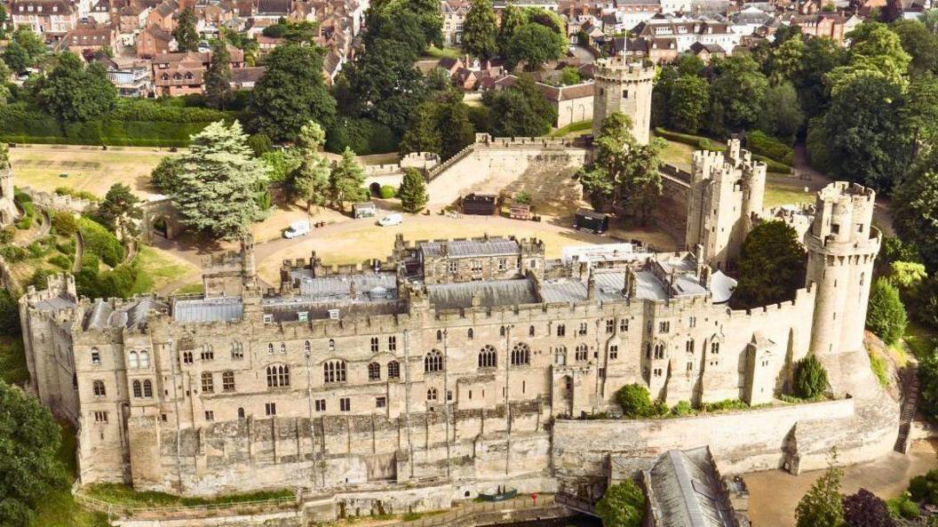 An aerial view of Warwick castle, which is light brown with multiple turrets. It has dozens of windows and a grass area in the middle of the walls.