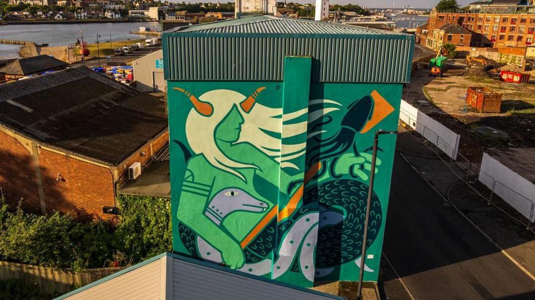 Giant mural covering the side wall of a high warehouse-type building. In shades of green, with some orange detail, it depicts a river god with flowing locks, horns and a fish tail, holding a creature which appears to be an eel.