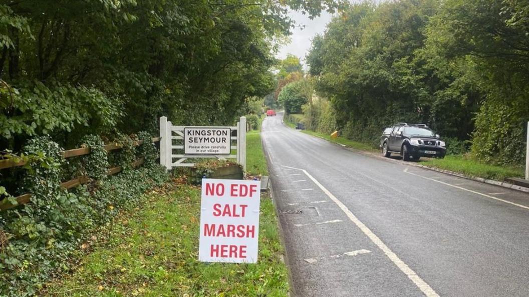 Rural road with trees surrounding it. There's a welcome to "Kingston Seymour" sign and a smaller sign on the ground saying "No EDF Salt Marsh here" in red capital letters