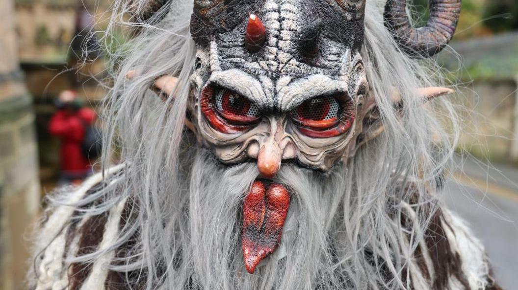 A person in a Krampus mask with white fur, horns, red eyes and a long tongue 