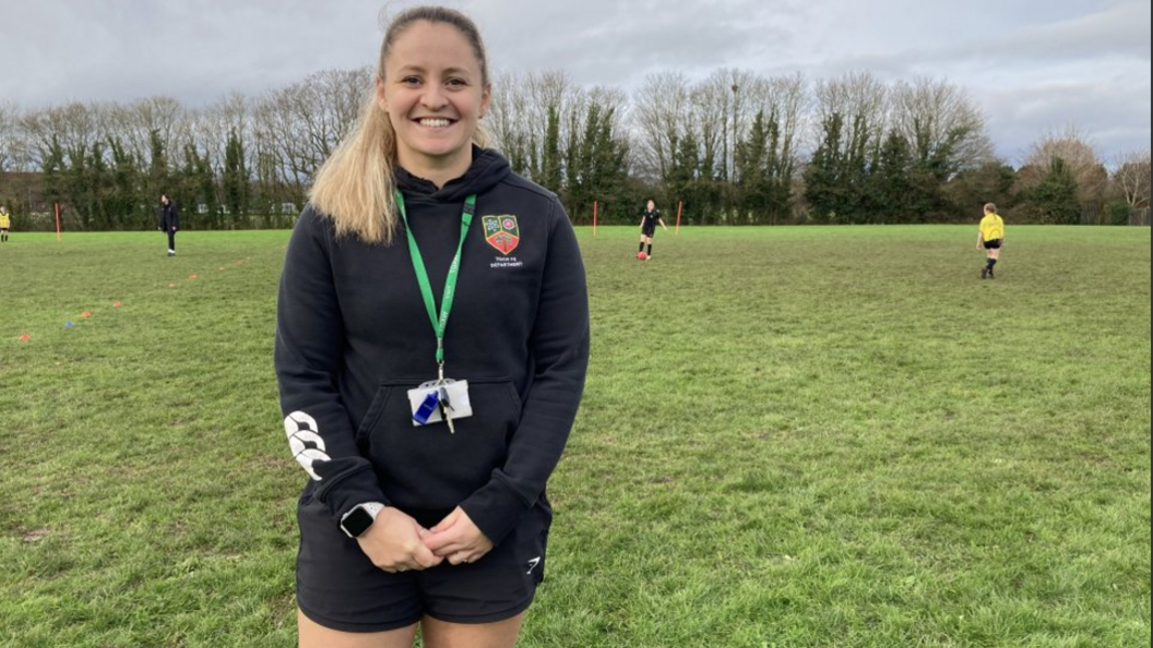 Bethany Marson with shoulder-length blond hair in a ponytail and dressed in a black hoodie with a school logo on the left hand side and black shorts. She is smiling on a football pitch