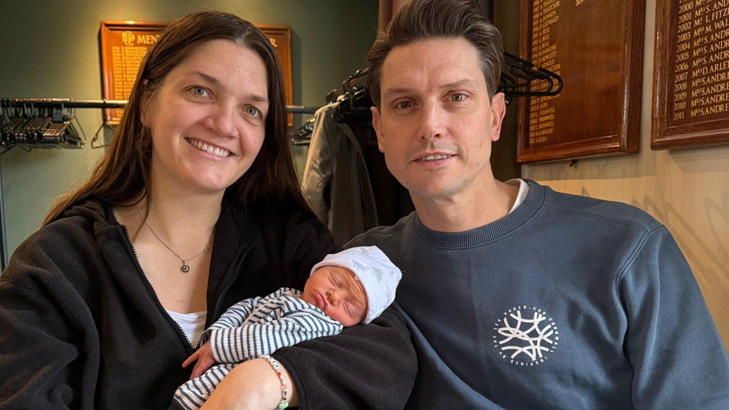 Lucy is holding a small baby boy, sitting next to her husband, Adam are both smiling at the camera. Lucy has dark hair, blue eyes and is wearing a black cardigan while Adam has dark hair and eyes and is wearing a blue sweatshirt.