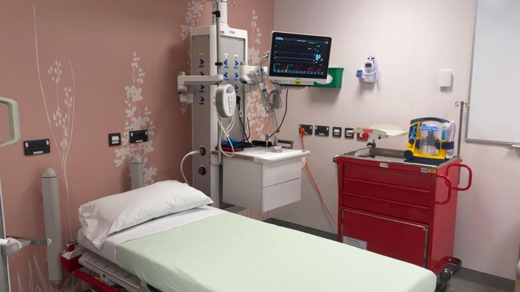 The pictures shows a cubicle on a ward. There's a bed in the middle, with an observation machine behind it, and a red cabinet to the side.