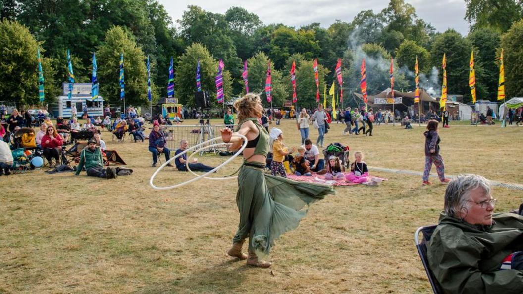 Woman with long blonde hair wearing a small green top and green trousers is holding a white hula hoop in each hand and dancing. There are people sitting on a ground sheet in the background and others sitting on the grass. There are flags and trees in the background, as well as food stalls.