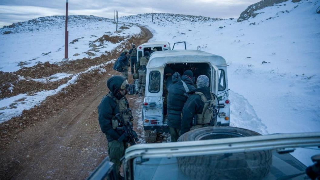 Israeli soldiers stand on top of a rocky hill with snow covering much of the ground, with soldiers loading or unloading items from the back of white vehicles 