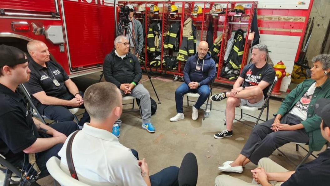 A group of people sit on plastic chairs in a circle. They are at a fire department. A fire engine/fire truck can be seen in the background