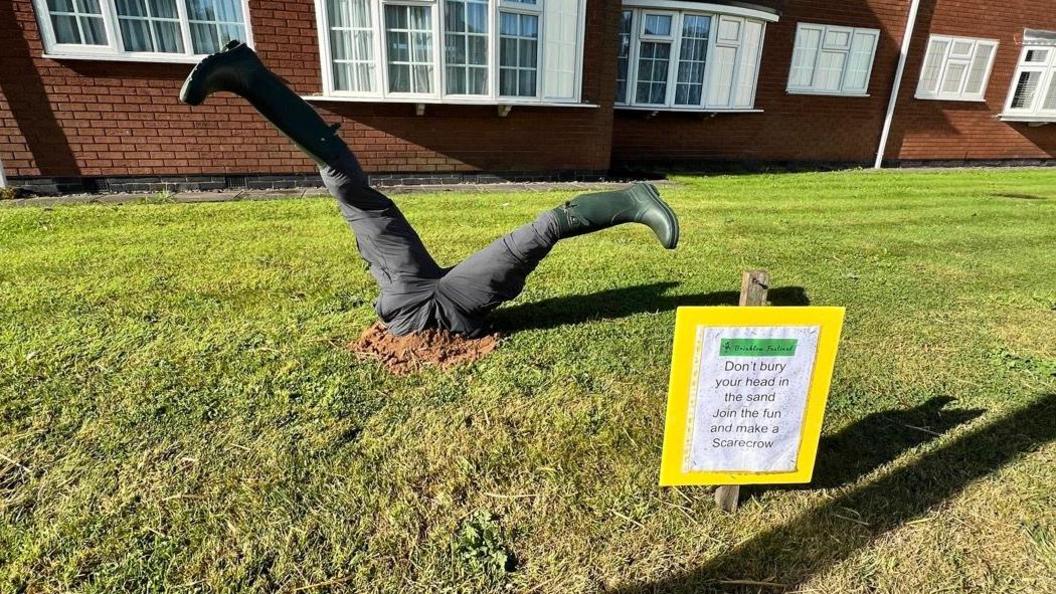 The legs of a scarecrow planted into grass. The scarecrow is dressed in black trousers and green wellies. A sign says 'Don't bury your head in the sand. Join the fun and make a scarecrow'.