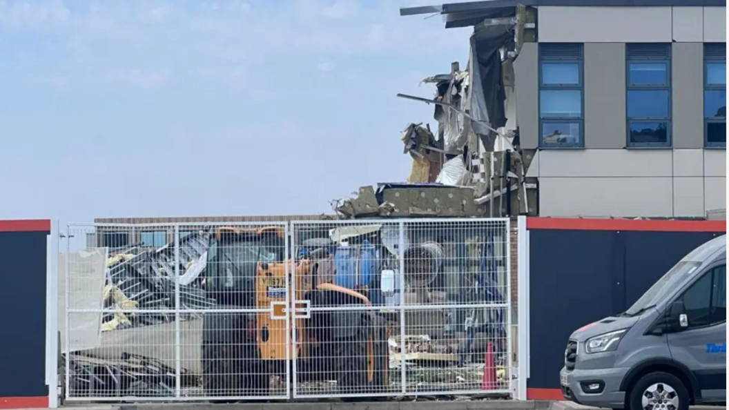 A building which has been partially demolished stands behind security fencing. A demolition vehicle is partly visible, along with a van on the nearside of the fence, which is positioned in a neighbouring car park.