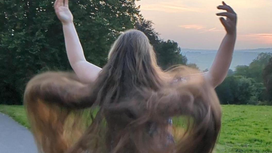 The back of Ruth Tripp with her arms in the air. She has long brown hair and is stood in front of a field with a sea view in the distance as the sun sets. 