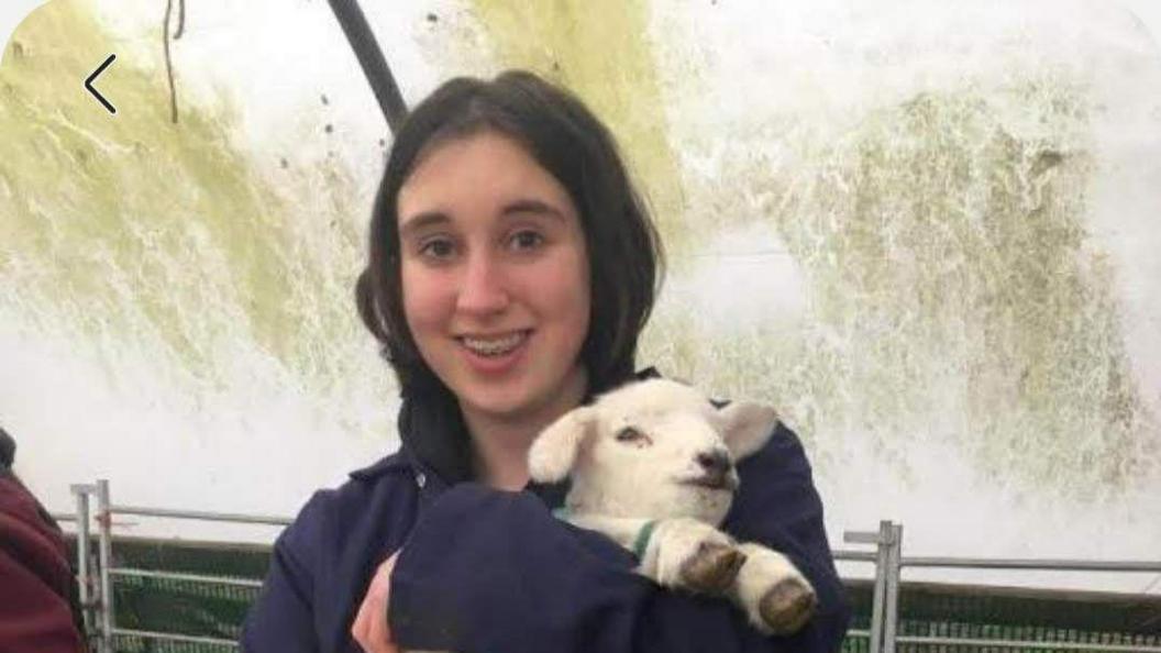 A teenage girl with brown hair and braces holds a newly-born lamb