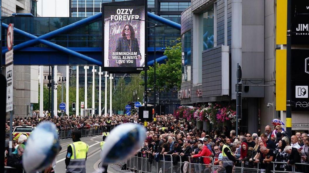 Thousands of people line either side of a road. Above them is a board that has a picture of Ozzy Osbourne on it. It reads "Ozzy forever, Birmingham will always love you"