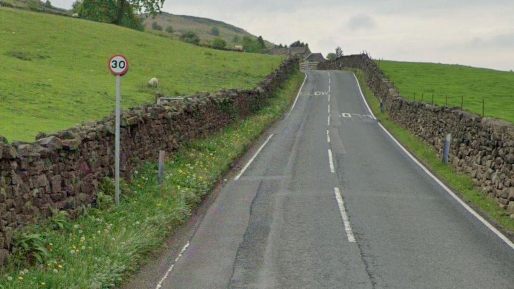 A country road runs between two fields, one of which has a sheep in. There is a 30mph sign at the side of the road.