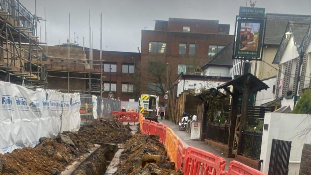 Road dug open opposite pub entrance with orange fencing around the building material.