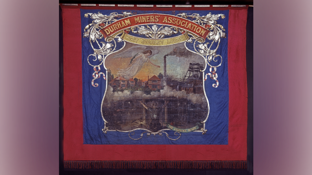 A red and royal blue banner that reads 'Durham Miners' Association, West Stanley Lodge'. An angel can be seen floating in the sky with houses in the background and a smoke tower and colliery. A person is holding their arms up to the sky underneath in darkness.
