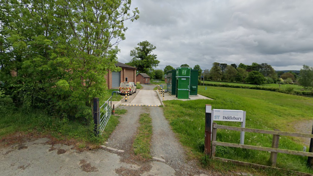 A brick building next to a small concrete road and green metallic structures behind a silver metal gate in a rural setting