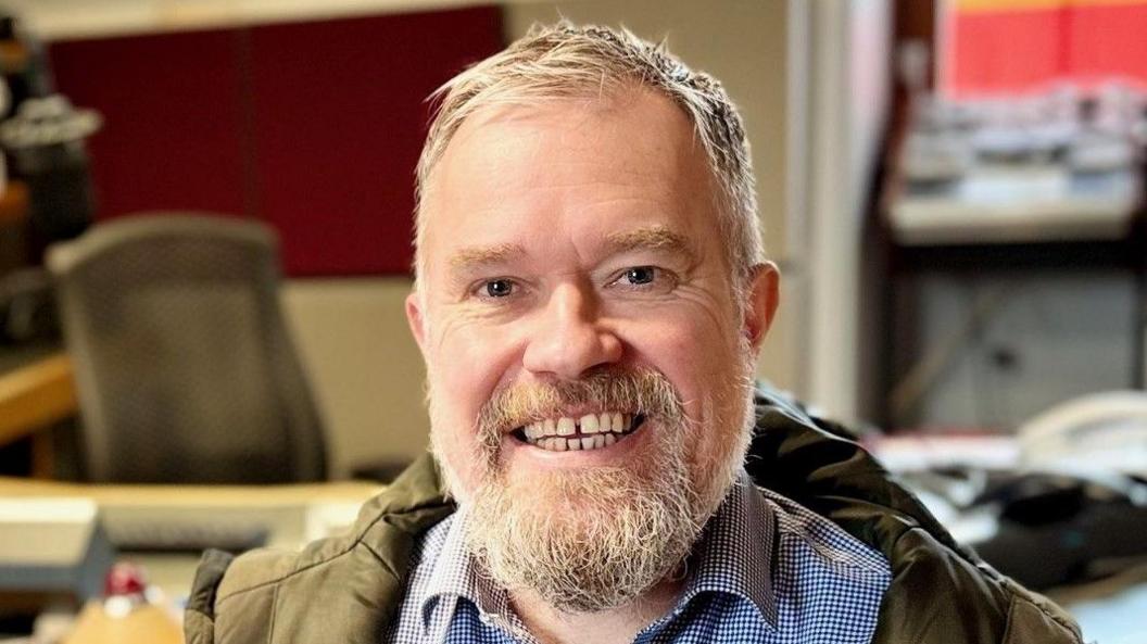 Paul Olvho, who has short, grey hair and a short, grey beard, smiles in a radio studio