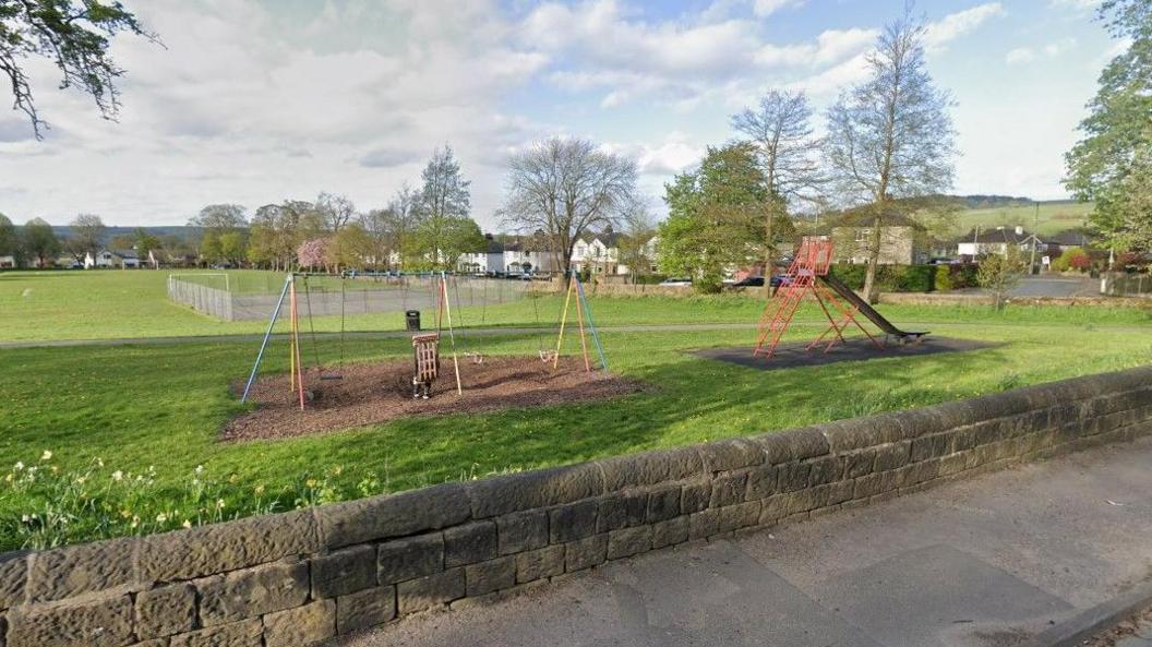 Menston Recreation Ground on a sunny day. Two sets of swings and a slide can be seen on a meadow. In the distance, rows of houses are dotted along the road.