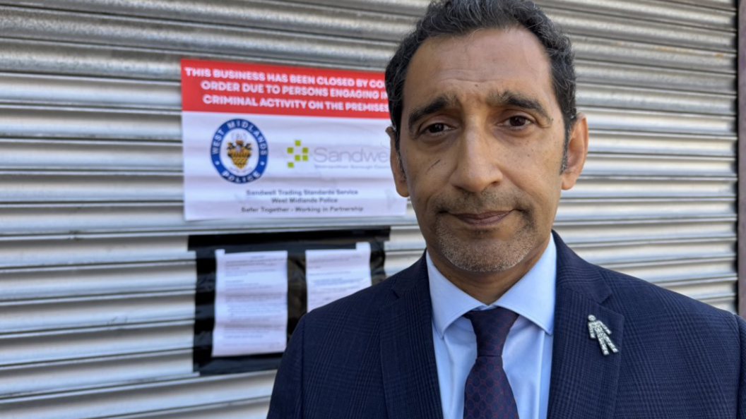 Shokat Lal is wearing a navy suit with a blue shirt and a blue tie stands in front of a closed shop premises. The silver shutters are down and a notice tells people it's closed. 